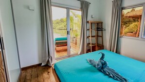 A bedroom with a large bed, a wooden floor, a window with curtains, and a view of a deck with a bench and plants. - Coral Reef Over Water Bungalow, Bedroom, King Bed, Living Room, Kitchenette (Bastimentos Island)