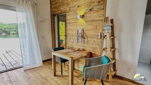 A room with a wooden wall, a table, a chair, and a view of a body of water through a window. - Coral Reef Over Water Bungalow, Bedroom, King Bed, Living Room, Kitchenette (Bastimentos Island)