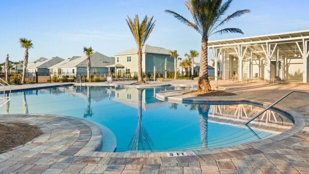 A swimming pool with a clear water surface, surrounded by a paved walkway, palm trees, and residential-style buildings in the background. - The Tipsy Turtle- Port St. Joe/Windmark Beach + Pet Friendly (Port St. Joe)
