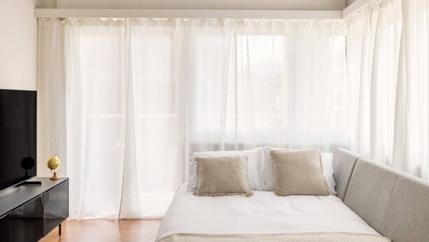 Un dormitorio con cama, televisión sobre un mueble negro y cortinas translúcidas.