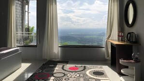 A room with a large window, a view of the countryside, a small table with a kettle, and a patterned rug. - Contana By Travelink (Srimulyo)