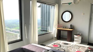 A hotel room with a bed, a window with curtains, a small table with a kettle, a mirror, and a view of the city. - Contana By Travelink (Srimulyo)