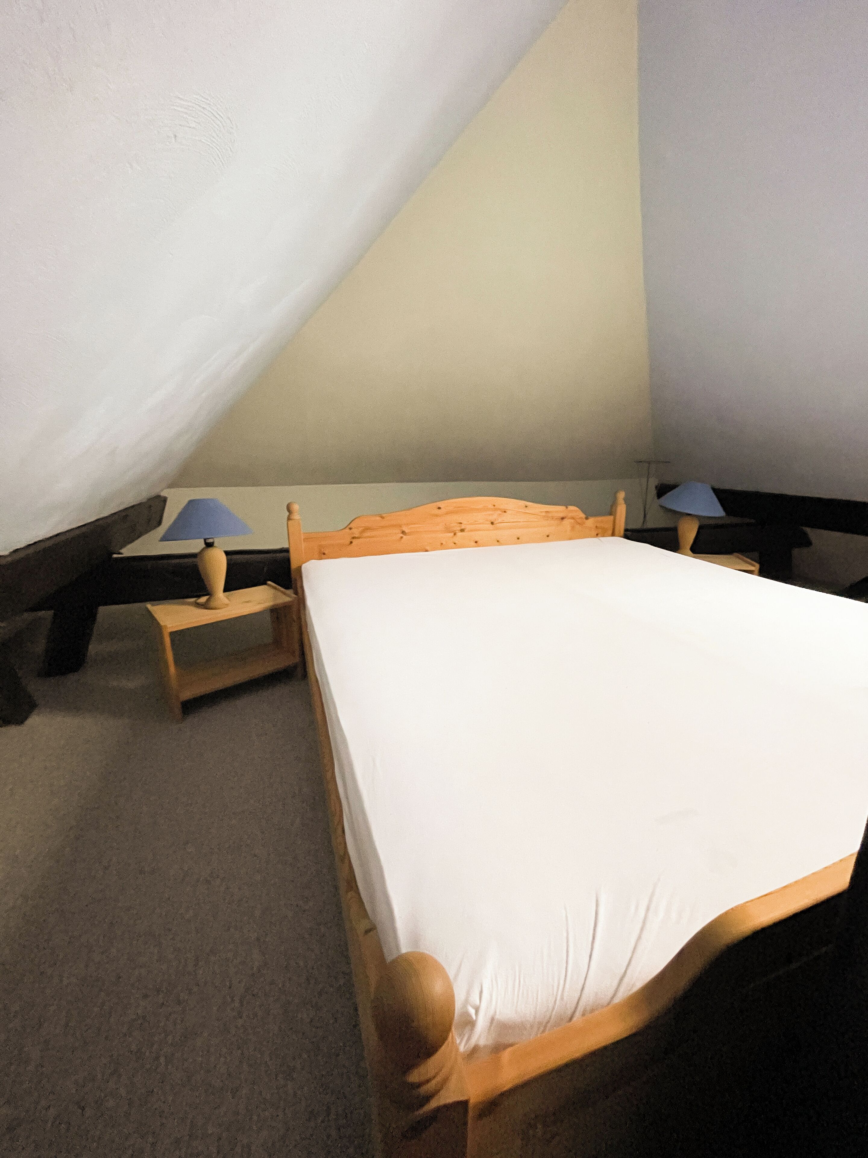 Una habitación pequeña y acogedora con una cama individual y dos mesitas de noche, cada una con una lámpara. La habitación tiene techo inclinado y paredes lisas.