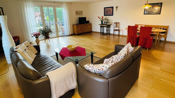 Ein Wohnzimmer mit einer Couch, einem Glastisch, einem Esstisch mit Stühlen und einer Küche im Hintergrund.