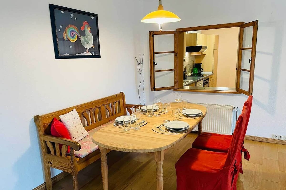 Ein Essbereich mit einem Holztisch, gedeckt für vier Personen, einem roten Stuhl, einer Holzbänkl mit Kissen und einem gerahmten Kunstwerk an der Wand.