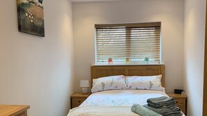 A neatly arranged bedroom with a wooden bed, a bedside table with a lamp, a small nightstand, a decorative rug, and a window with blinds. - Cozy barn conversion within minutes from Silverstone. (Towcester)