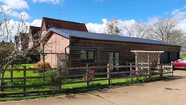 A rustic log cabin with a metal roof, surrounded by a wooden fence and a paved driveway. - Cozy barn conversion within minutes from Silverstone. (Towcester)
