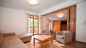 Ein geräumiges Wohnzimmer mit einer Couch, Sesseln, einem Holztisch und einem großen Fenster mit Vorhängen.