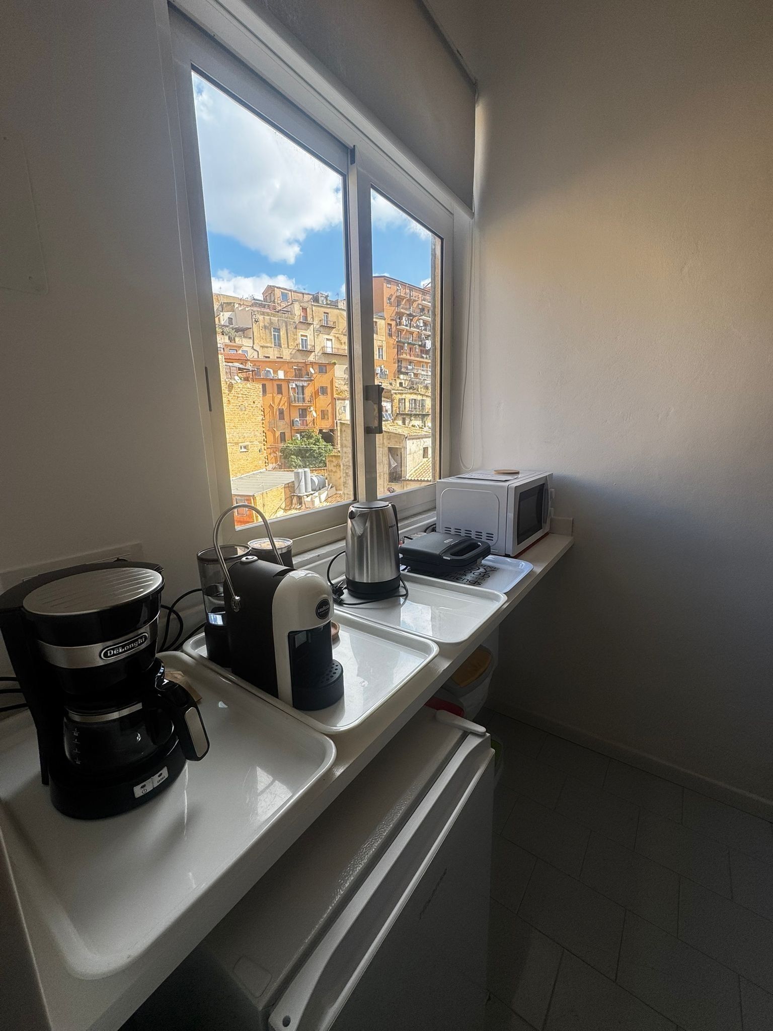 Une cuisine équipée d’une machine à café, d’une bouilloire et d’un grille-pain sur le plan de travail, avec une fenêtre donnant sur un paysage urbain.