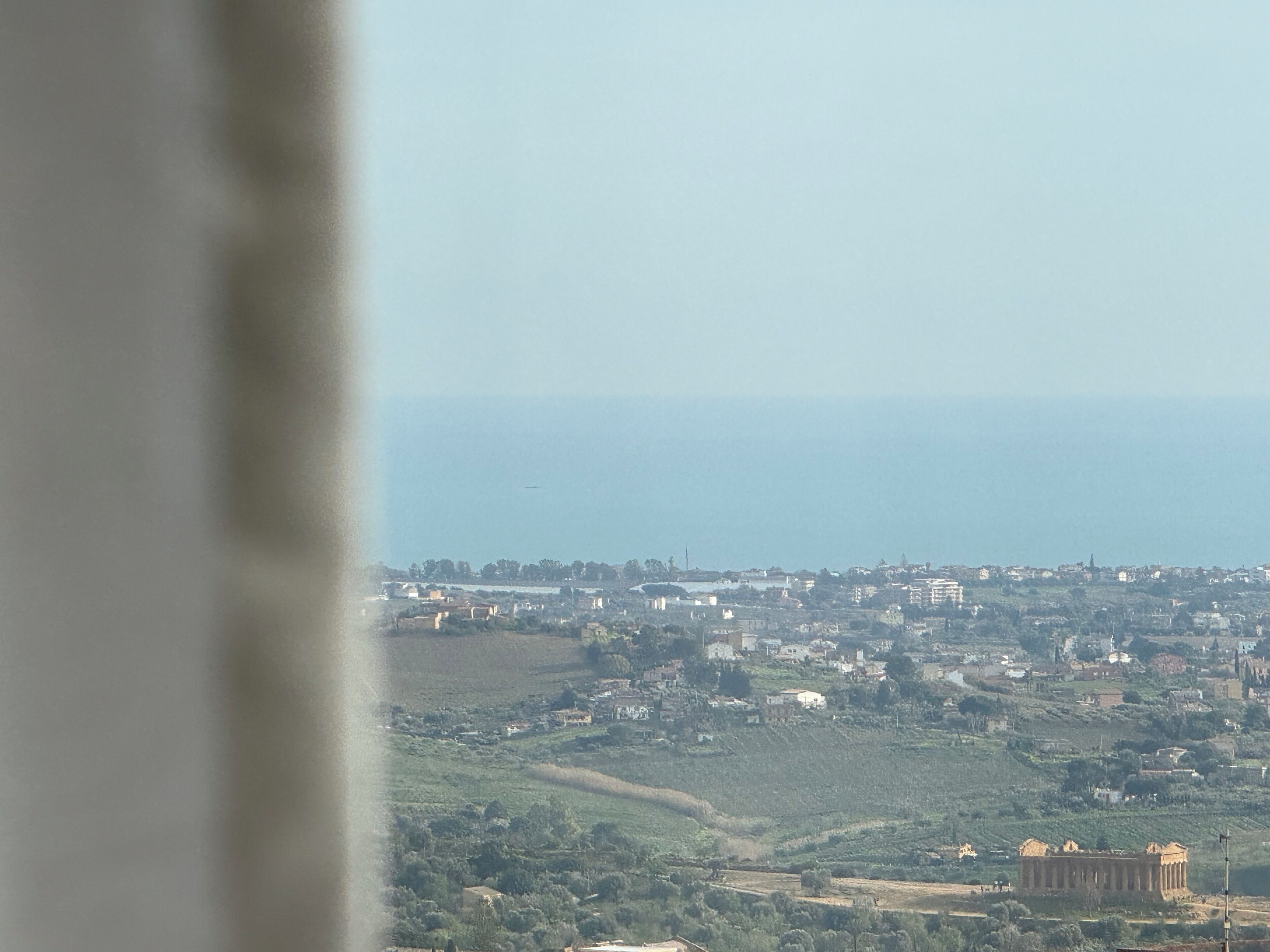 A coastal town with a prominent ancient temple, a clear sky, and a distant airplane.