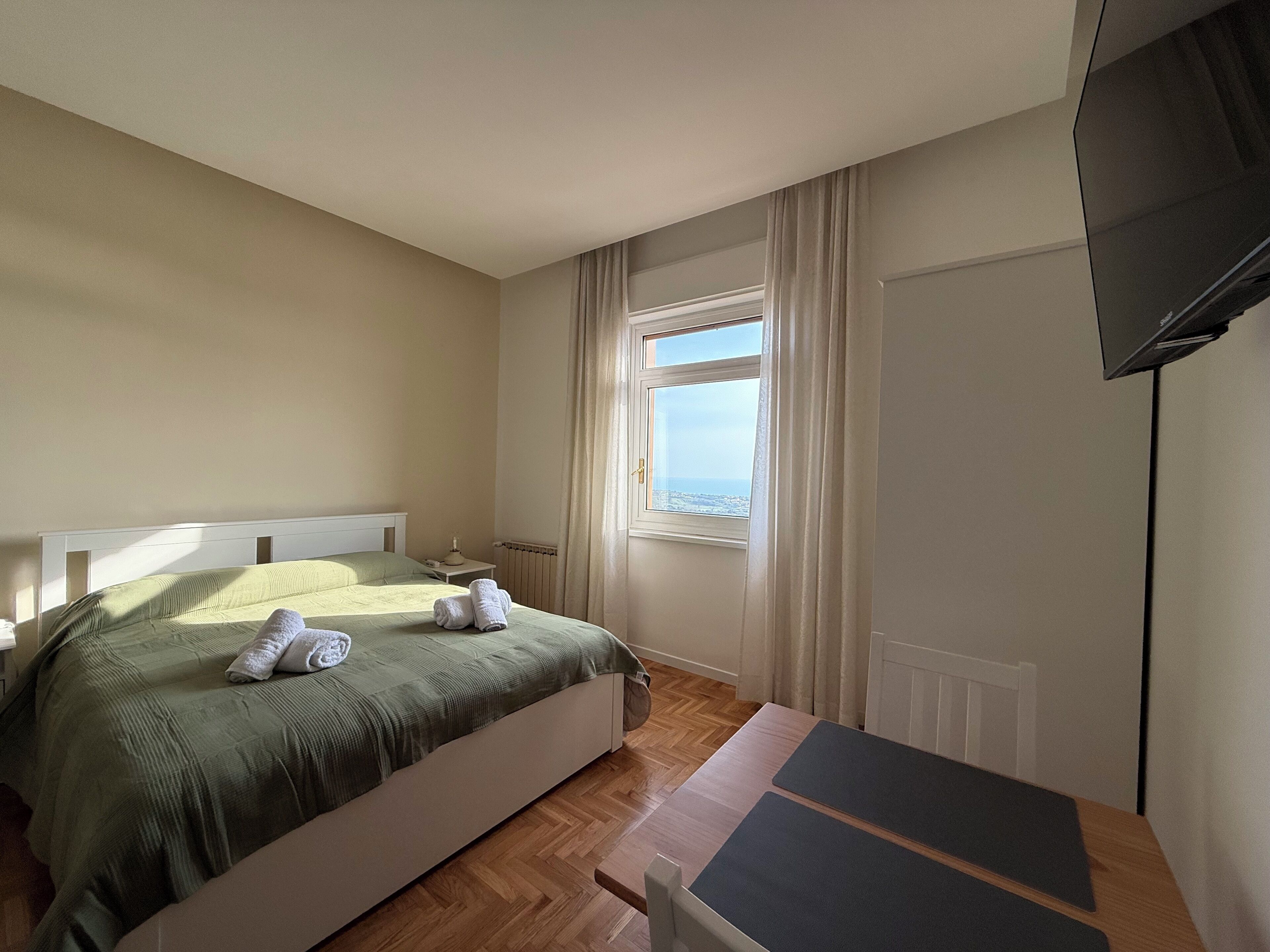 A bedroom with a bed, a nightstand, a window with curtains, a radiator, and a television mounted on the wall.