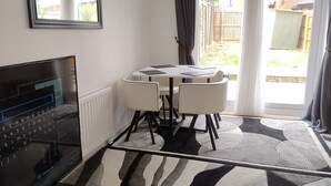 Ein Zimmer mit Fernseher, der eine Tastatur zeigt, einem Esstisch mit Stühlen und Blick in einen Garten durch eine Glastür.