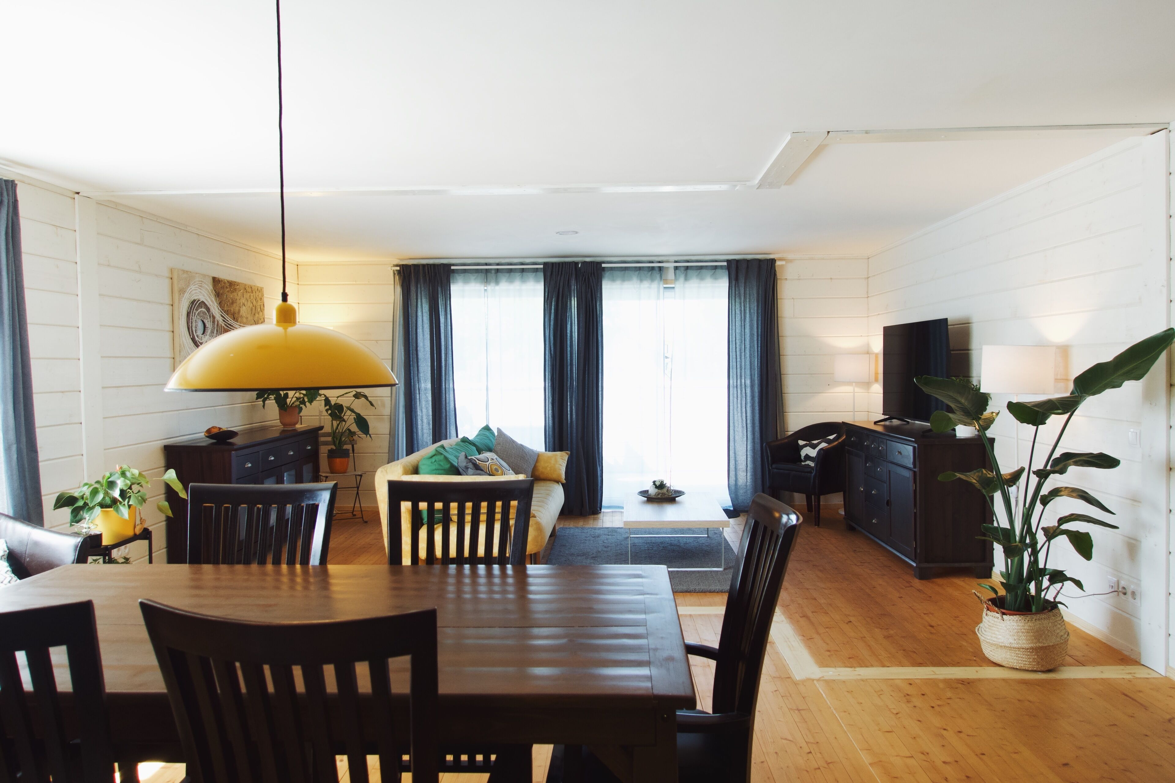 Un salon moderne avec une table à manger en bois, des canapés foncés, un téléviseur et une grande fenêtre avec des rideaux.