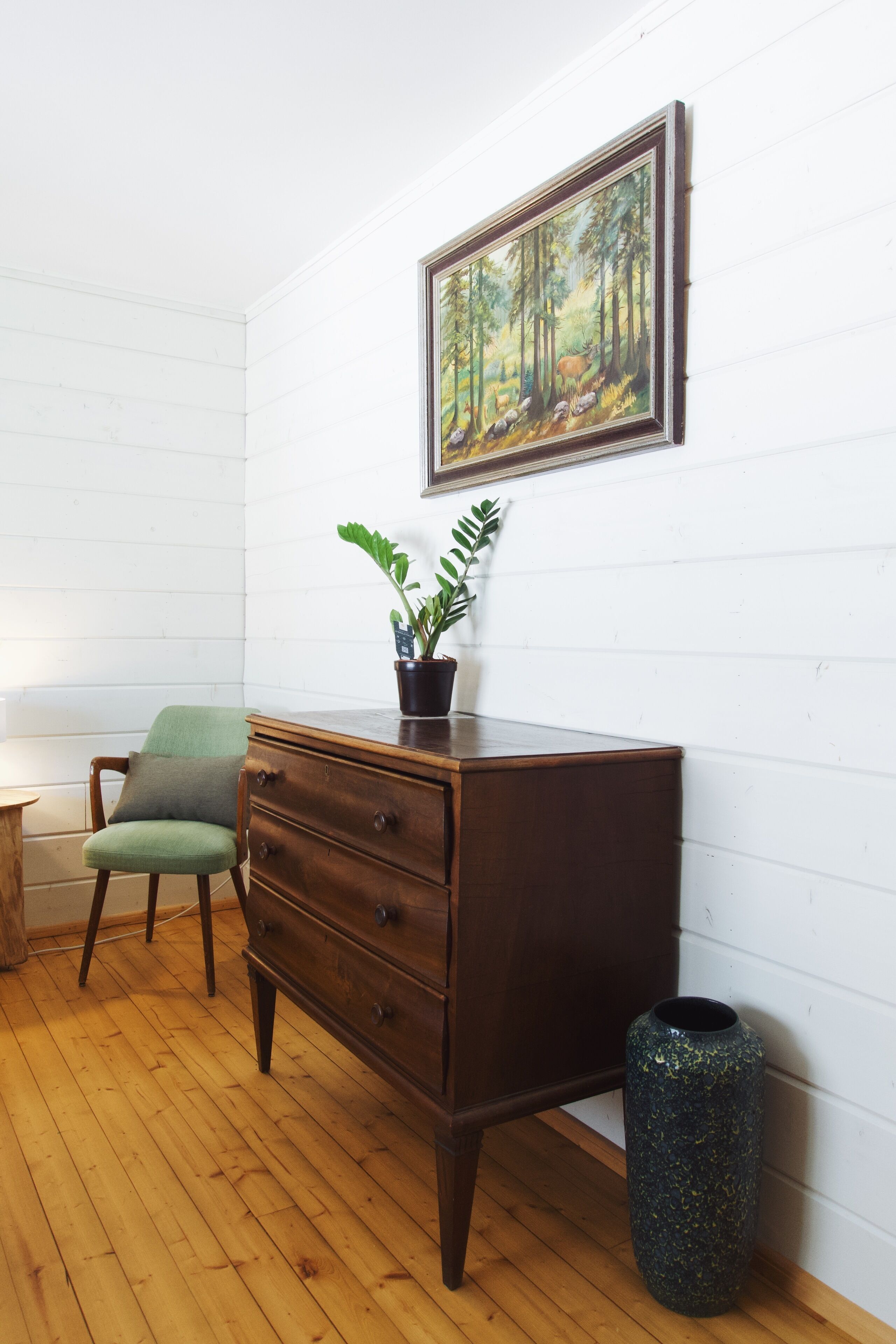A room with a wooden floor, a green chair, a dark wooden dresser, a potted plant, a framed painting of a forest, and a trash can.