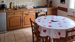 A kitchen with wooden cabinets, a tiled backsplash, a dining table with a floral tablecloth, and a window with a teddy bear decoration. - Quiet and surrounded by nature 10 mm from the A75 freeway (Montcel)