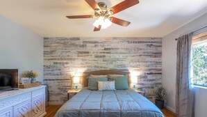 Une chambre à coucher avec une tête de lit en bois, un lit recouvert d’une couette bleue, un ventilateur de plafond et une table de chevet avec une lampe et une plante.