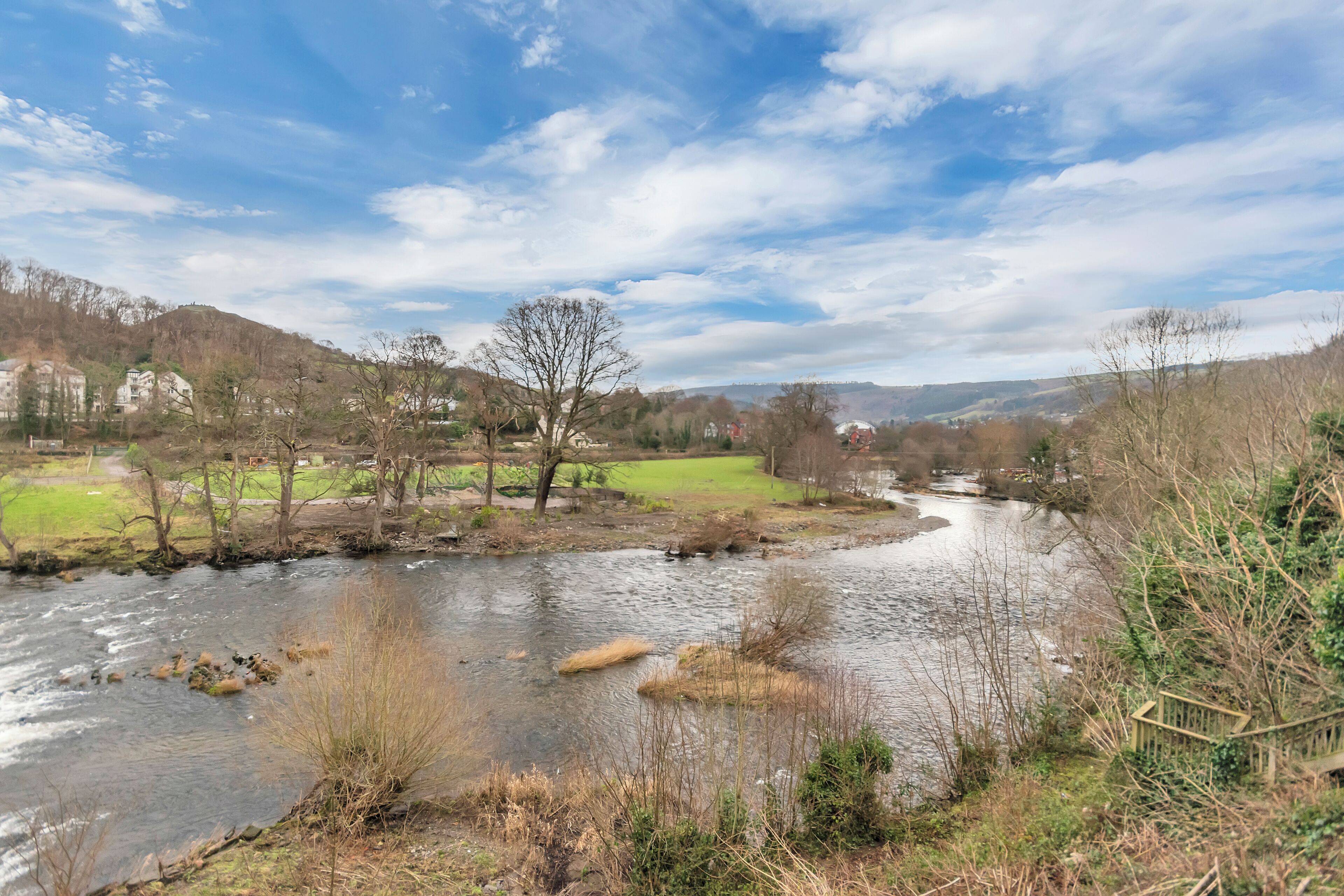 A river flowing through a rural landscape with houses and a grassy field in the background.
