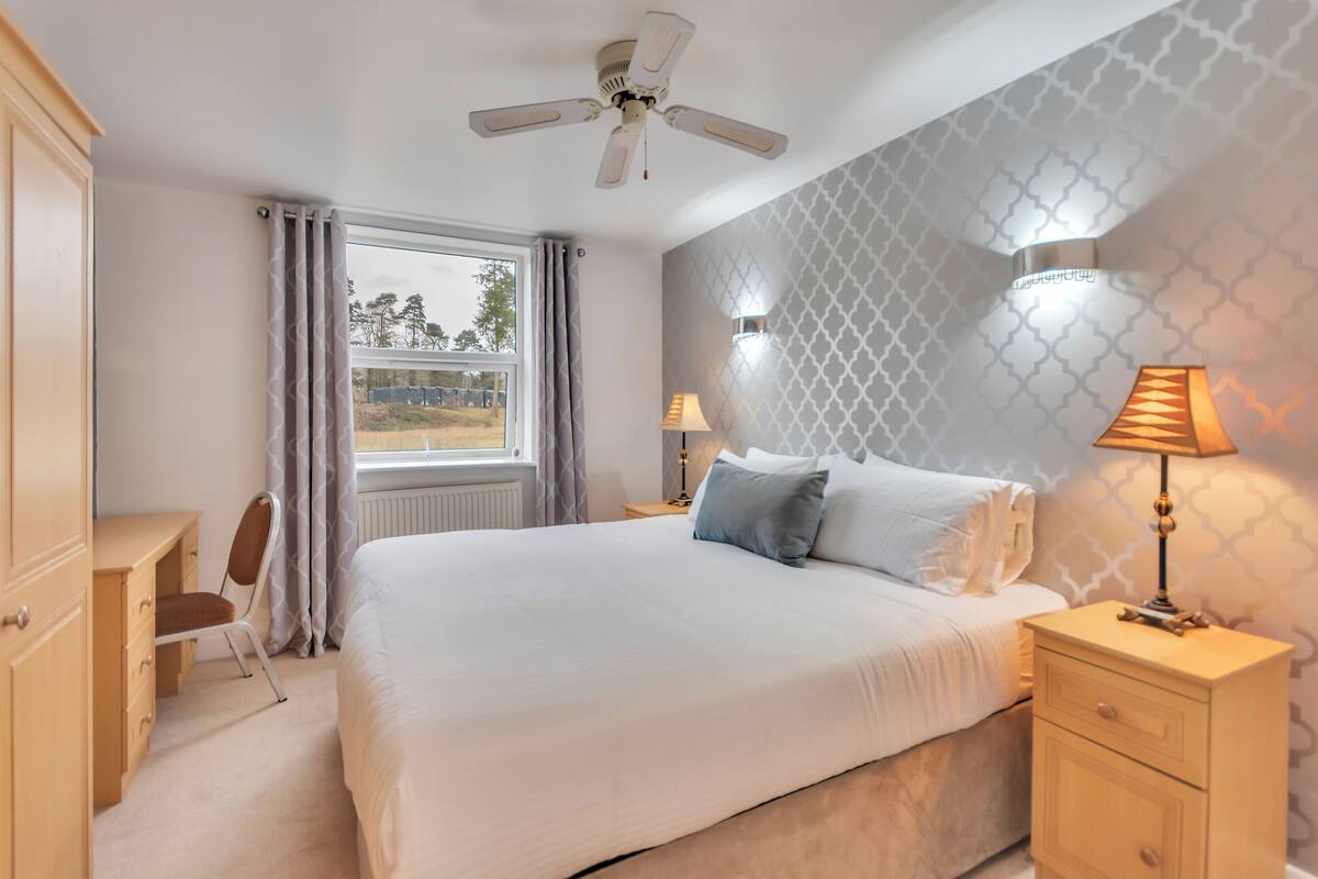 A bedroom with a bed, a desk, a chair, a ceiling fan, and a window with curtains.