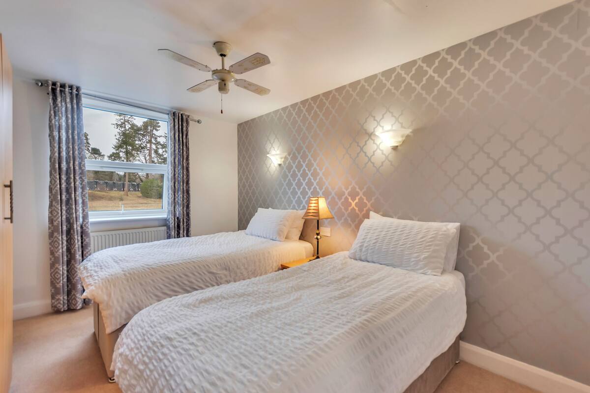 A bedroom with two beds, a ceiling fan, a window with curtains, and patterned wallpaper.