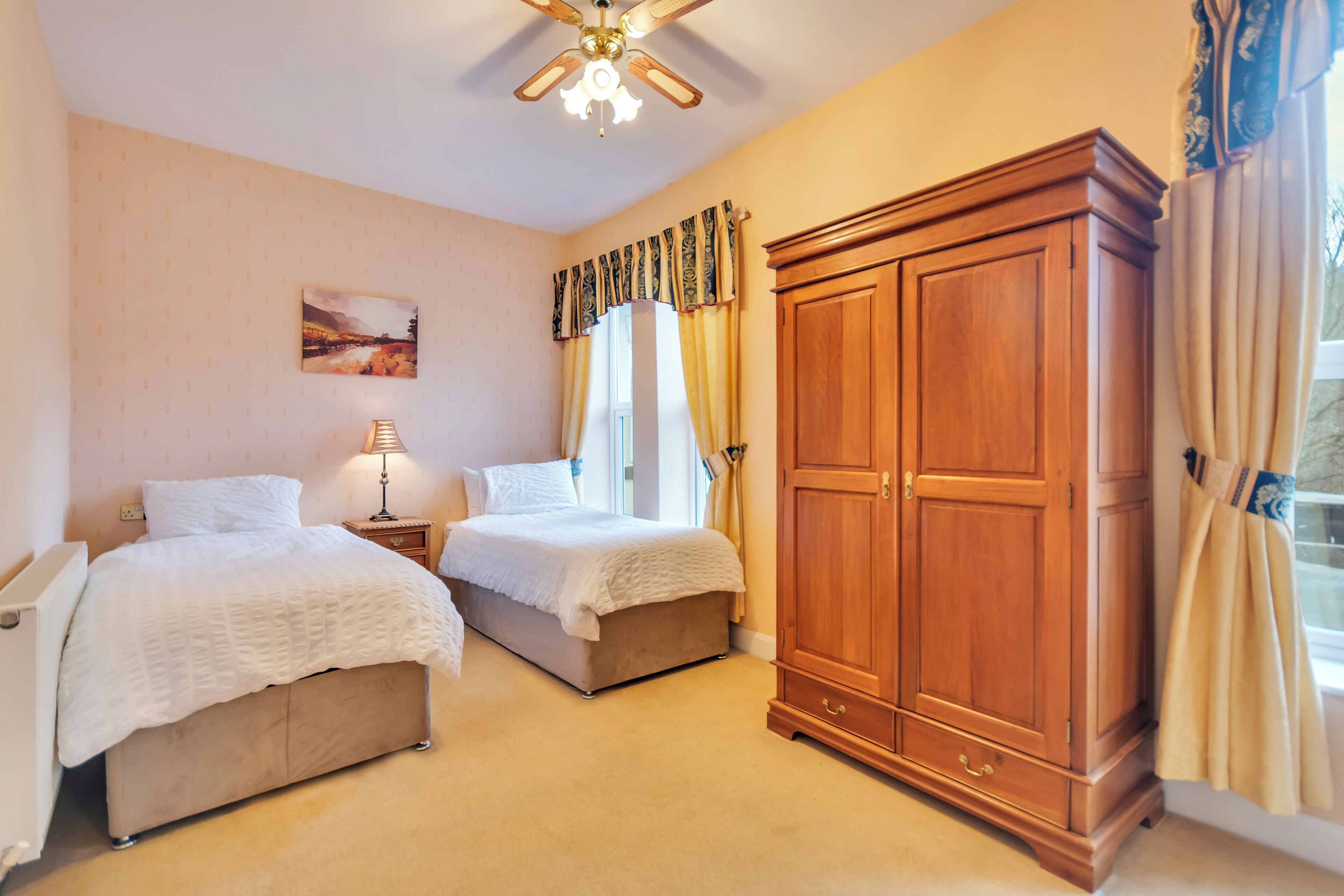 A bedroom with two beds, a wooden wardrobe, a ceiling fan, and a window with curtains.