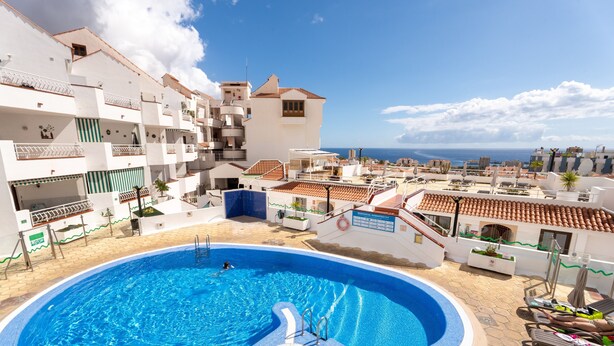 A swimming pool surrounded by a white building with balconies, a clear sky, and a view of the sea.