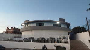 A modern building with a curved facade, multiple windows, and a fenced parking area with several cars. - Corporate Accommodation (Parque das Universidades Campinas)