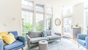 Een moderne woonkamer met een grijze bank, een blauwe fauteuil en een klein voetenbankje. Grote ramen zorgen voor natuurlijk licht en uitzicht op groen.
