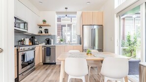 Een moderne keuken met roestvrijstalen apparatuur, een eethoek met witte stoelen en uitzicht op groen.