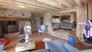 A rustic interior with exposed wooden beams, a stone fireplace, a kitchen area, and a seating area with a table and chairs.