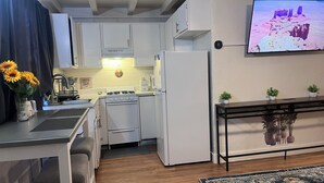 Een kleine keuken met witte kastjes, een fornuis en een koelkast. Er is een eethoek met een tafel en stoelen, en een aan de muur bevestigde televisie waarop een landschap te zien is.