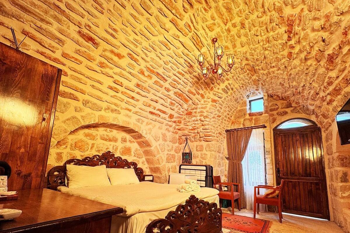 A room with a stone vaulted ceiling, a bed with a wooden headboard, a wooden dresser, a small table, and a chair.