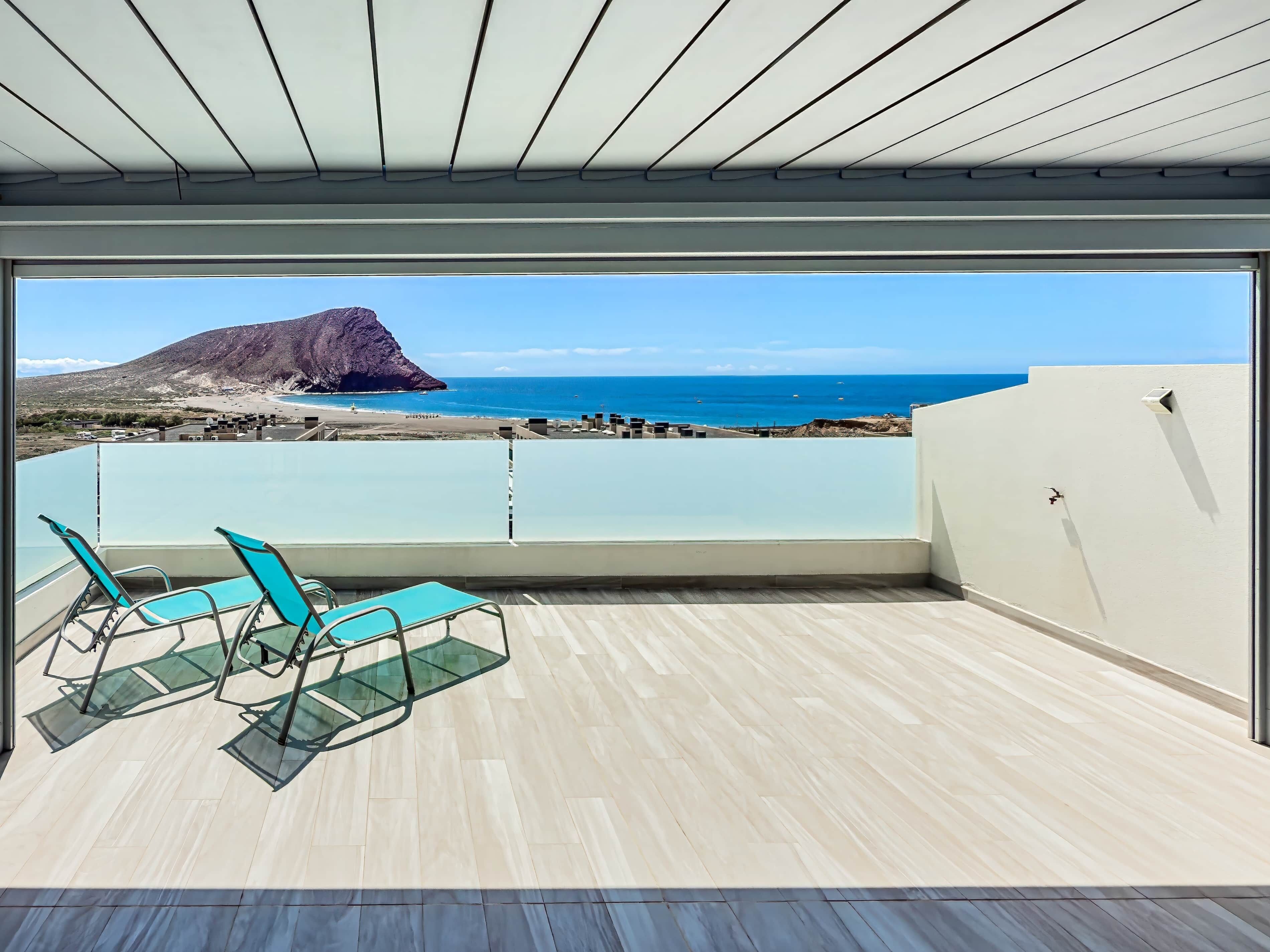 Een balkon met uitzicht op zee en een berg, voorzien van twee stoelen en een glazen tafel.