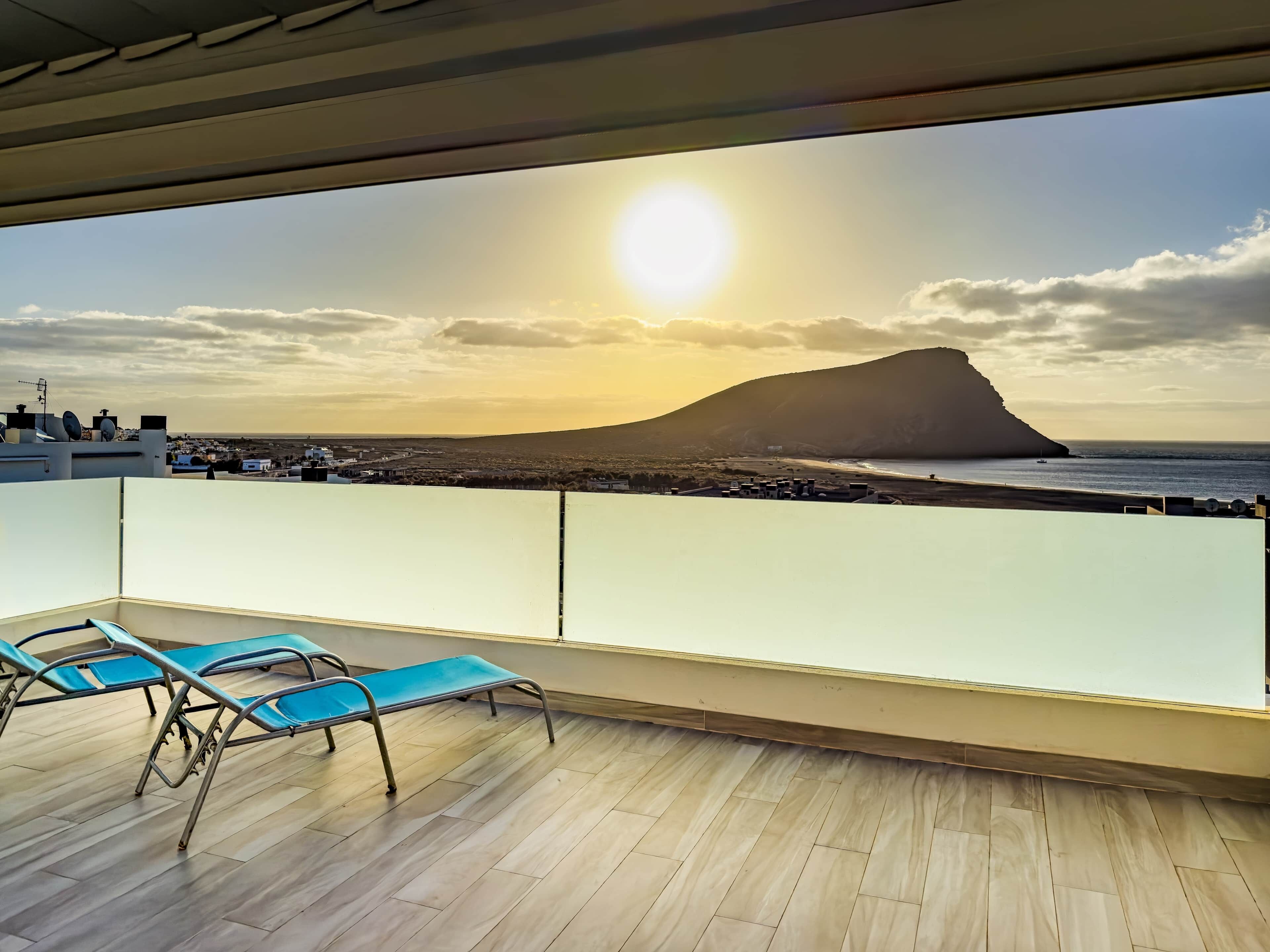 Een balkon met twee blauwe stoelen en uitzicht op zee en een berg bij zonsondergang.