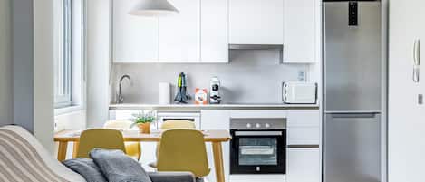 Une cuisine moderne avec des meubles blancs, un réfrigérateur en acier inoxydable, un four encastré et un coin repas comprenant une table et des chaises.