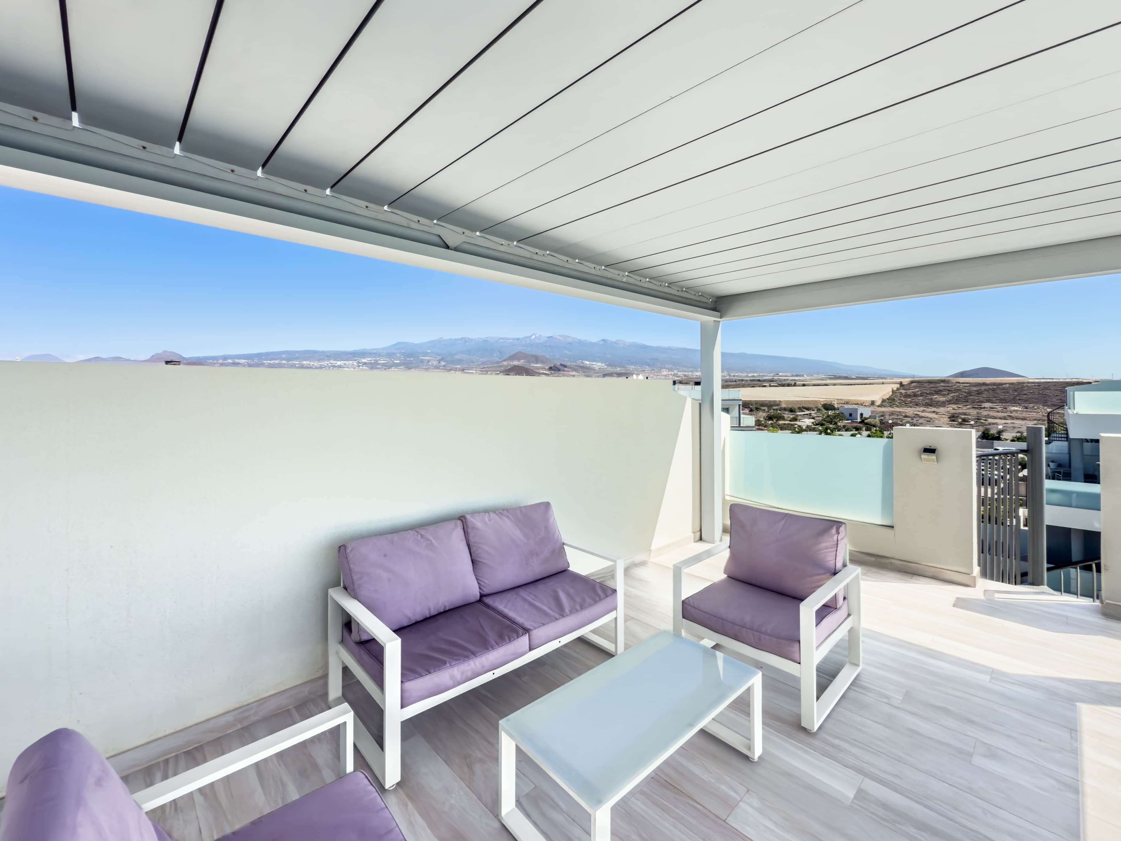 Un balcón con sofá y sillones, una mesa y vistas a montañas distantes.