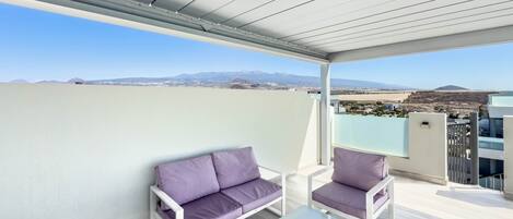 Un balcón con sofá y sillones, una mesa y vistas a montañas distantes.
