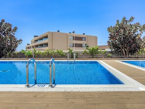 A swimming pool with a wooden deck, surrounded by a fence and trees, with a modern building in the background.