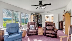 A living room with a blue armchair, two brown leather chairs, a ceiling fan, and large windows with a view of the outdoors. - 3BR Retreat in Prosser Perfect for Wine Lovers (Prosser)