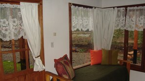 A cozy room with a wooden bed, a bench, and a view of a garden through large windows with lace curtains.