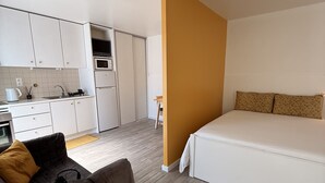 Een moderne hotelkamer met een kitchenette, een bed, een bureau met stoel, en een kleine tafel met lamp en boek.