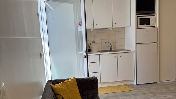 Een compacte leefruimte met een kitchenette, een bank met een geel kussen en een tafel met een lamp.
