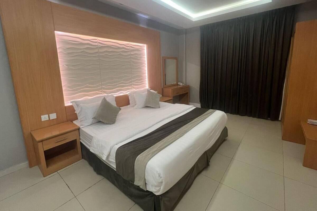 A modern hotel room with a large bed, wooden nightstands, a bedside table, and a headboard with built-in lighting.
