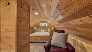 Ein gemütliches Schlafzimmer im Dachgeschoss mit schrägverkleidetem Holzboden, einem Bett mit Kissen, einem Sessel und einem Fenster.