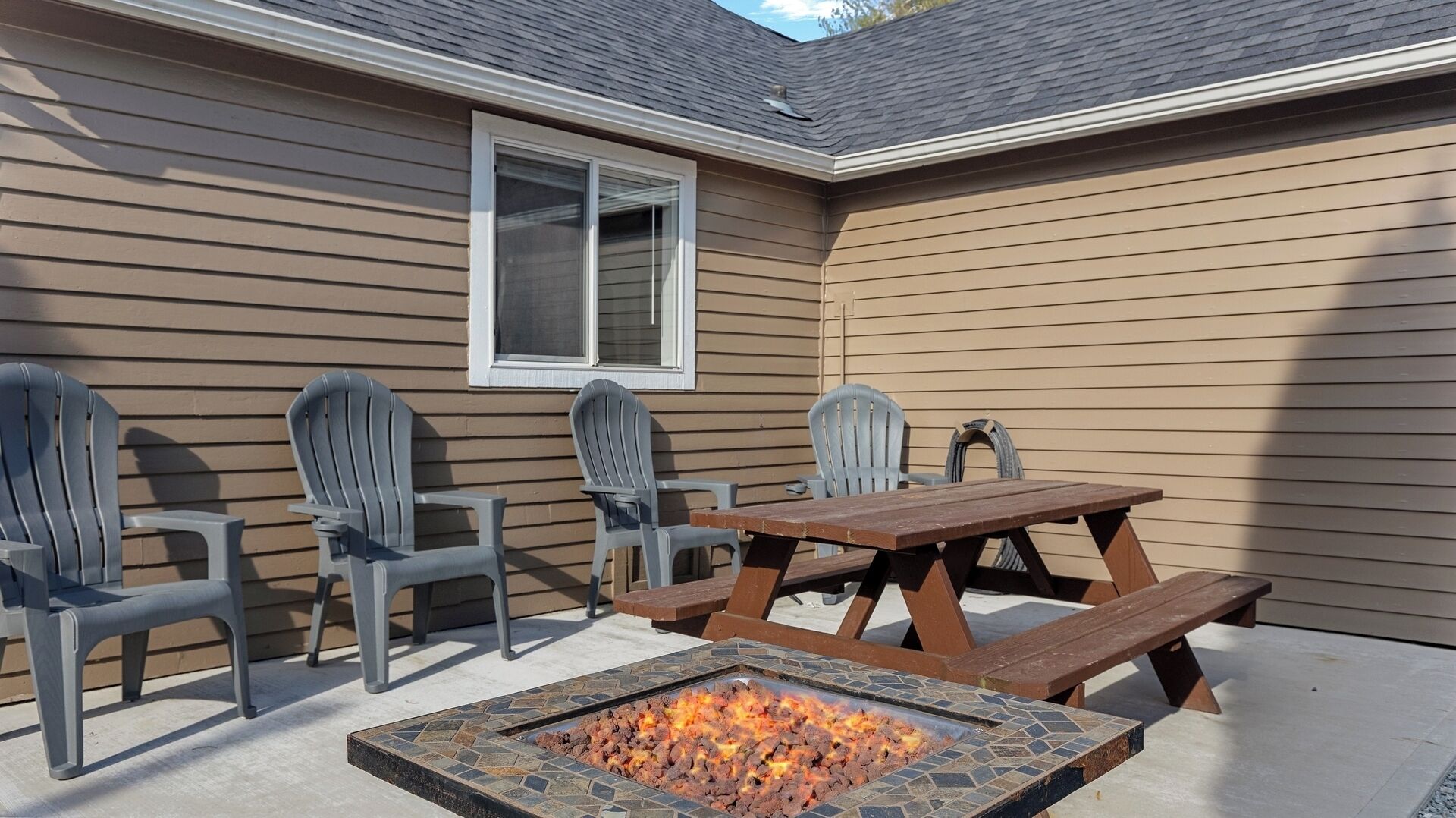 An outdoor patio with a fire pit, a wooden table, and four Adirondack chairs.