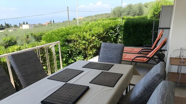 A terrace with a table set for four, surrounded by grey cushioned chairs, overlooking a lush green landscape with buildings in the distance. - Vacation apartment Lake Garda Cavaion Bardolino with pool (Cavaion)