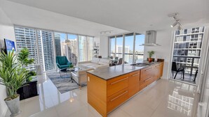 A modern kitchen with a large island, a dining area with a view of the city, and a living room with a sofa and a teal chair. - Sun, Style & Skyline | Modern Icon Brickell 2BR (Miami)