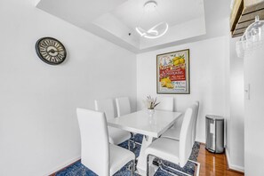 A dining area with a white table and chairs, a vintage clock, a poster, and a trash bin.