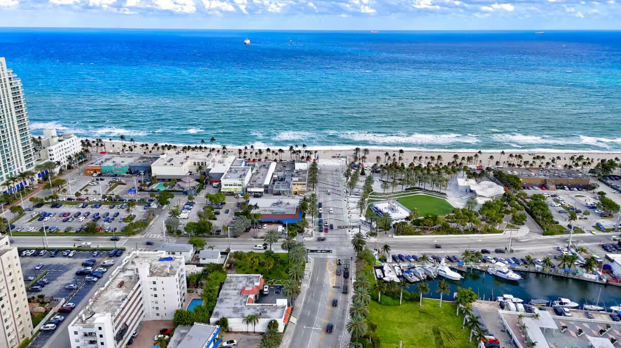 An aerial view of a coastal city with a marina, parking areas, and buildings along the beachfront.