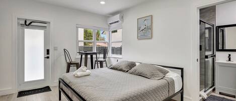A bedroom with a bed, a small table, and a chair. There is a window with a view of the outside.