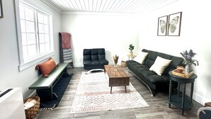 A living room with a wooden floor, a black sofa, a black armchair, a wooden coffee table, a bench, a patterned rug, and a window with blinds.